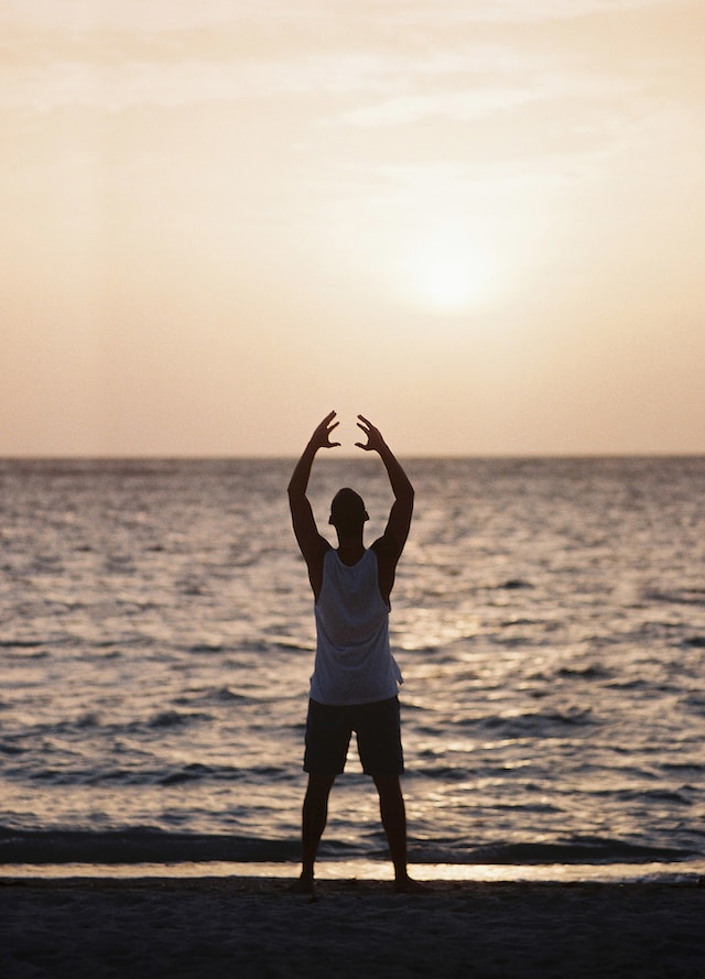 A man stretches his arms up while facing the sea.