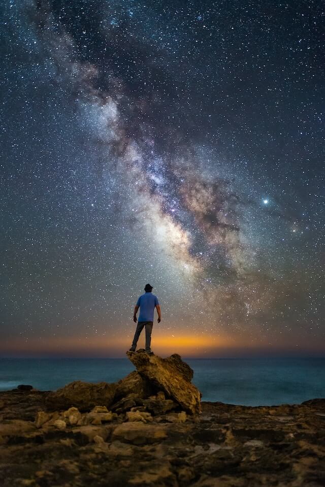 A guy stands in a jagged piece of rock while staring at the Milky Way.