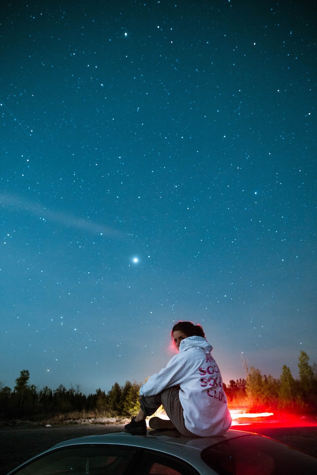 Woman sitting on top of her car, the starlit night sky in the background