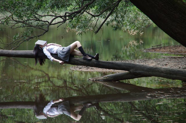 Girl with a book covering her face laying stretched out on a tree trunk over a still lake